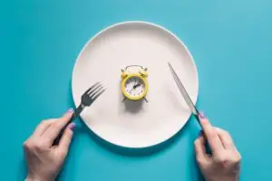 Plato vacío con un pequeño reloj despertador amarillo en el centro,flanqueado por un tenedor y un cuchillo simbolizando el control del tiempo en el ayuno intermitente.